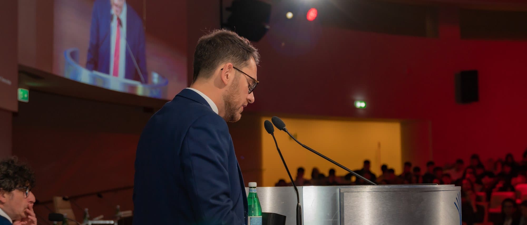 Kledis Brahimi parla da un leggio davanti a una sala piena, la sua immagine proiettata sullo schermo alle spalle
