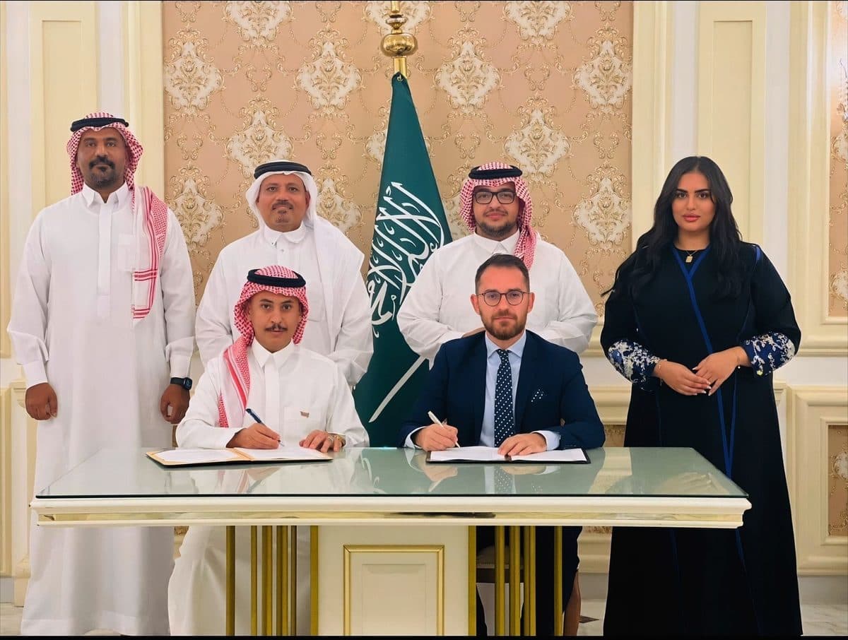 Two signatories at a table under the Saudi flag, with four people standing behind them.