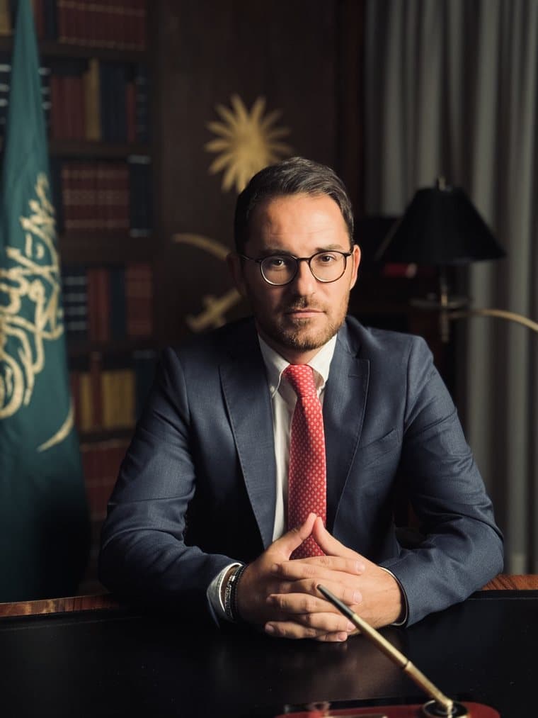Kledis Brahimi seduto a una scrivania, alle spalle una bandiera saudita e una libreria
