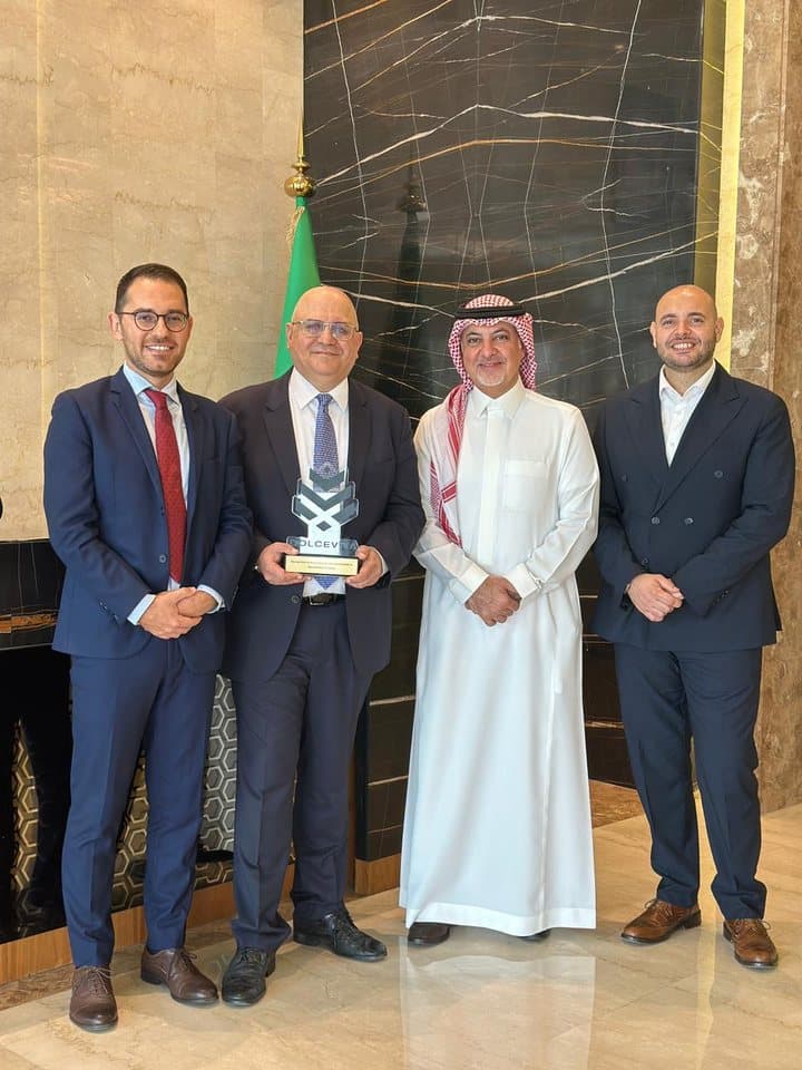 Quattro uomini nella hall di un albergo, uno con in mano un trofeo DolceVita, alle spalle una bandiera saudita
